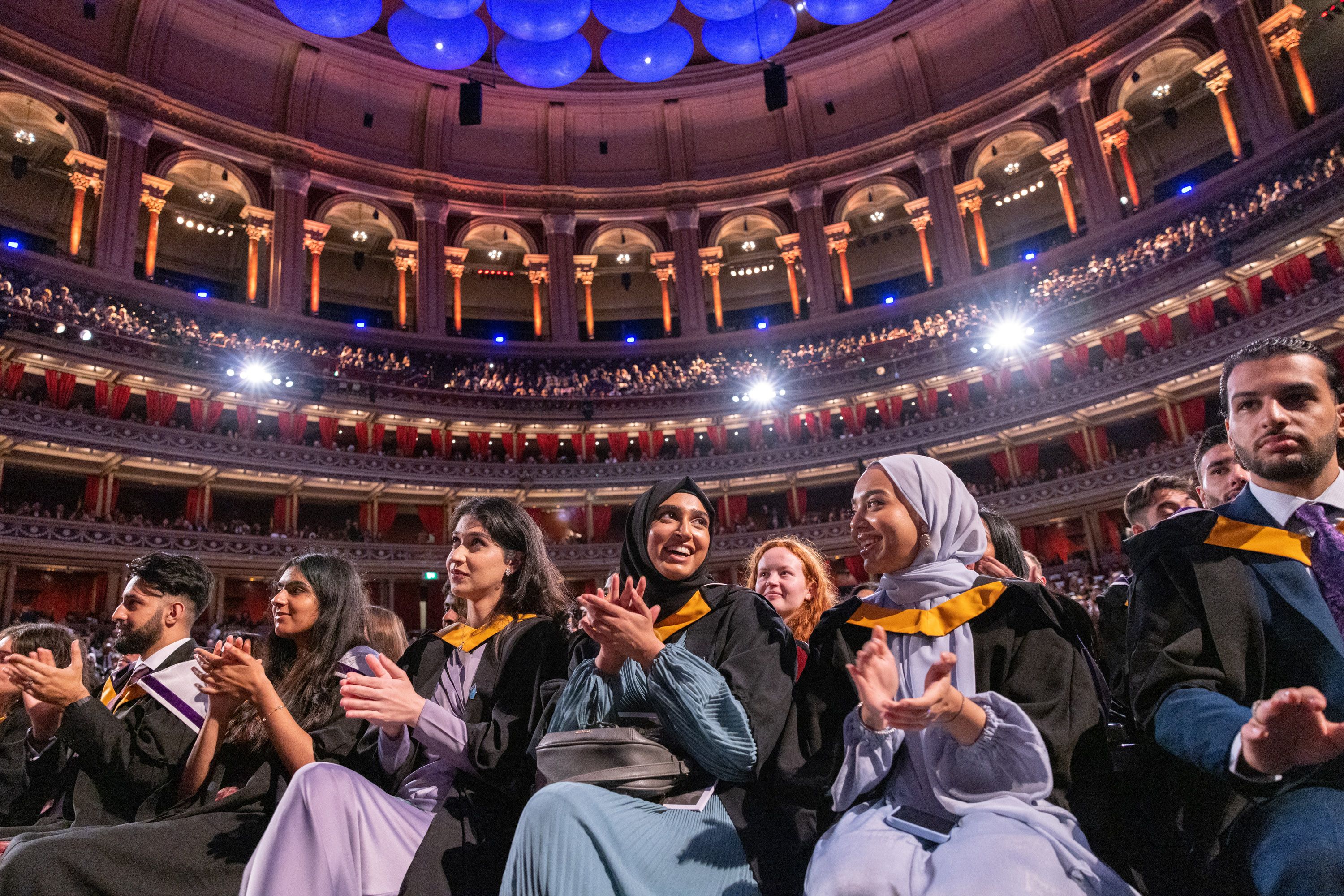 A pair of smiling students in headscarves applaud – behind them are the circles of the Albert Hall, all full