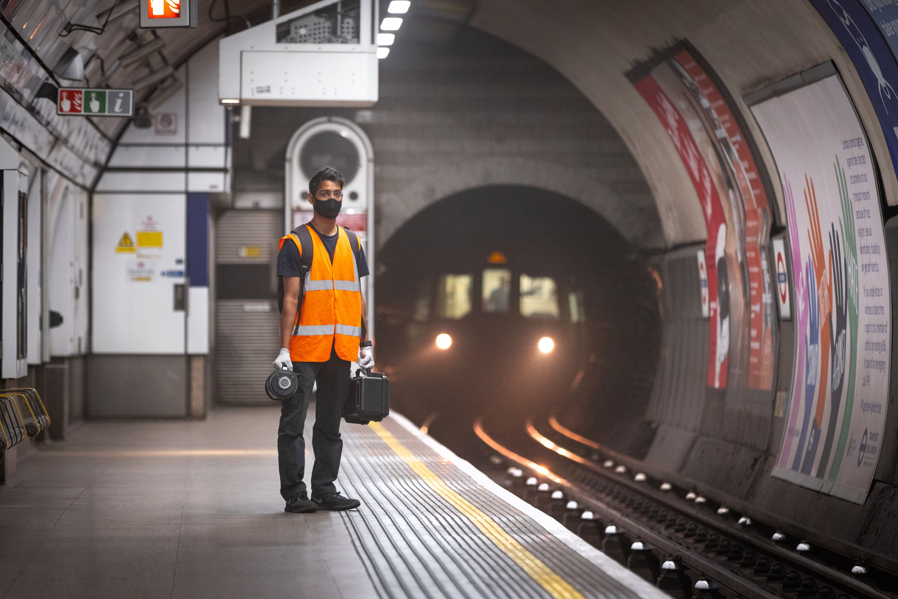 A student in a mask and hi-vis jacket stands on a tube platform while behind him in the tunnel we see the lights of an approaching train
