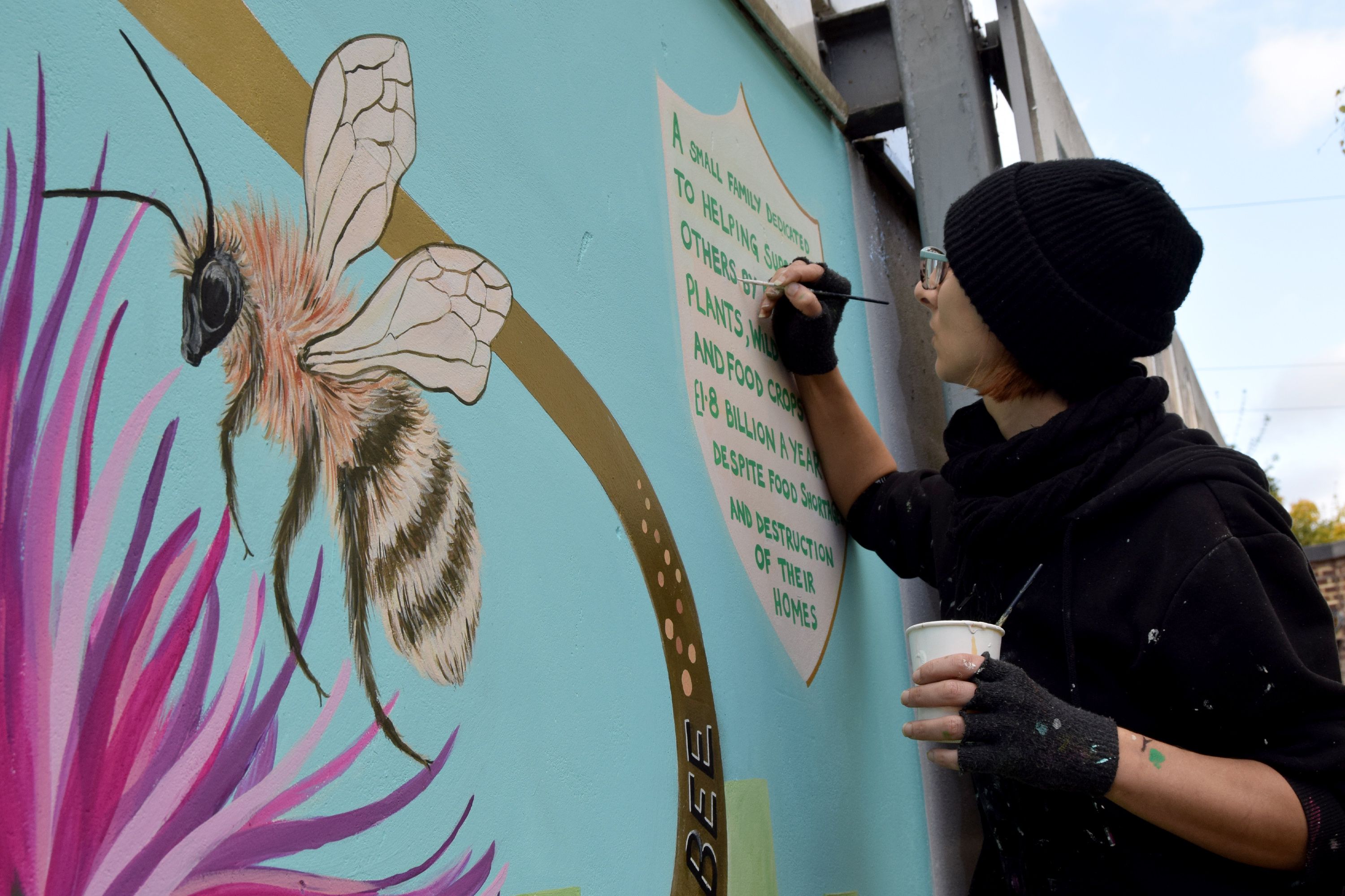 An artist in black wool hat and cutoff gloves painting a mural which includes a large bee