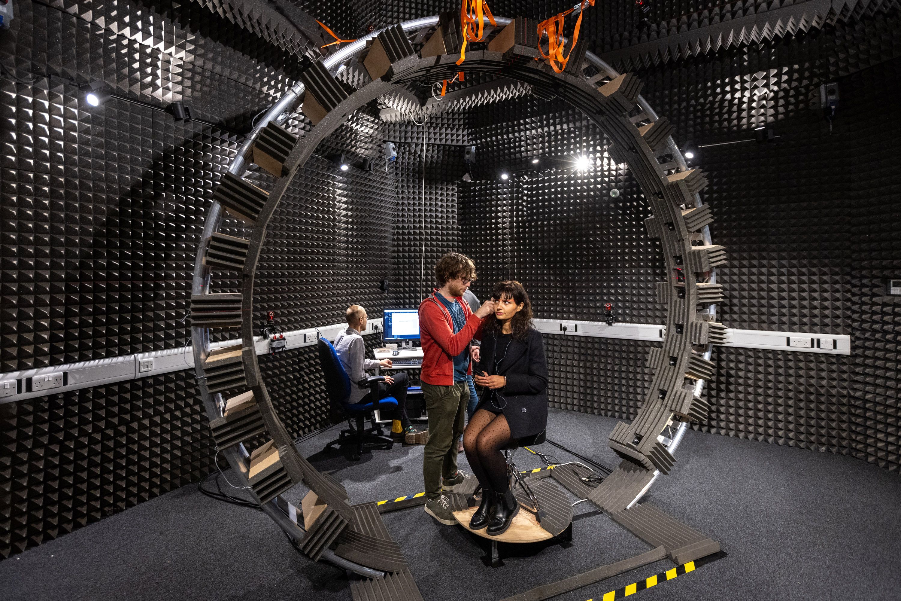 A woman sitting in a soundproof room in the middle of a huge metal loop, having headphones fitted to her by a researcher