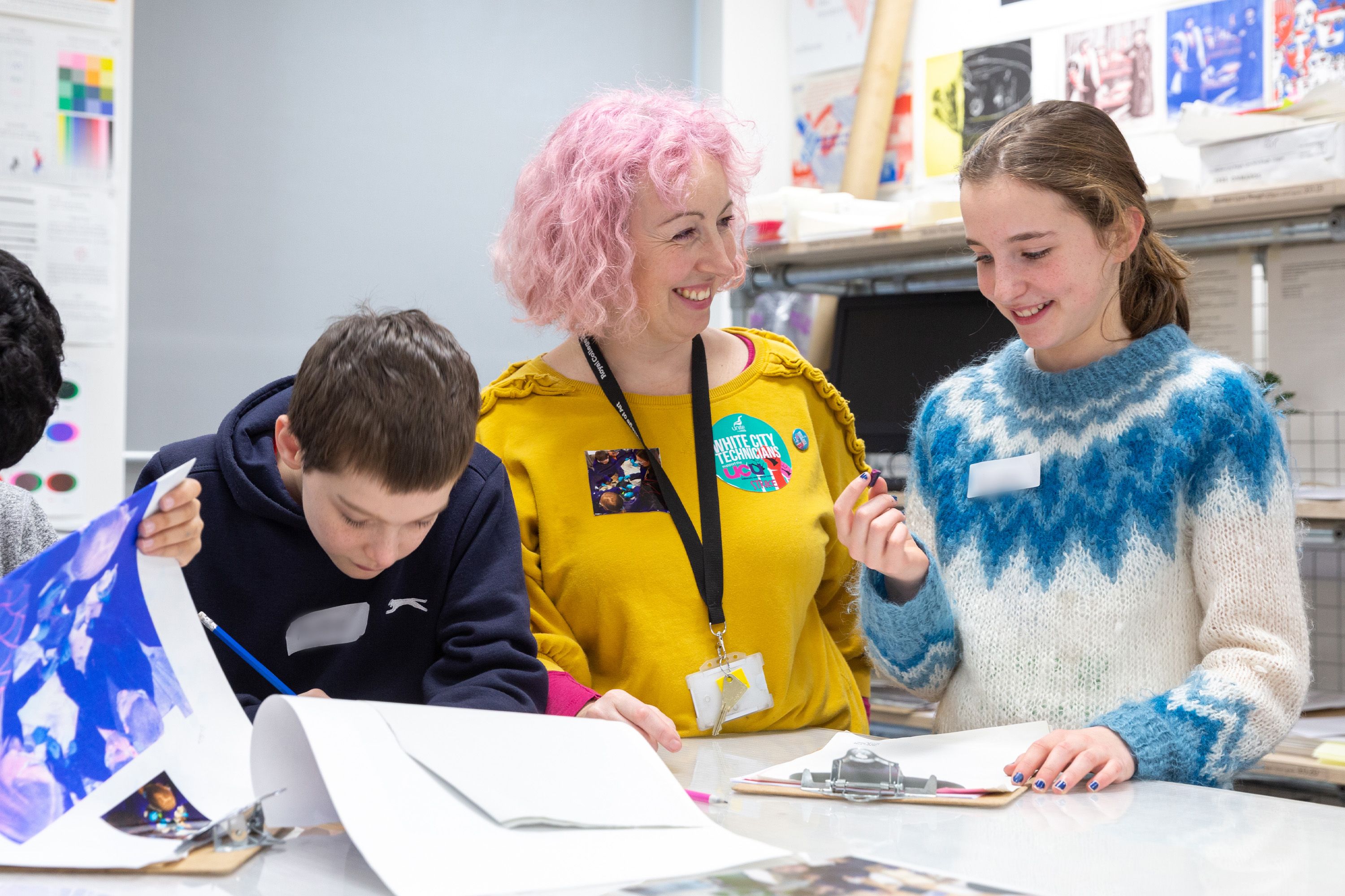 A pink-haired woman in a yellow top smiling at two children who are writing on paper