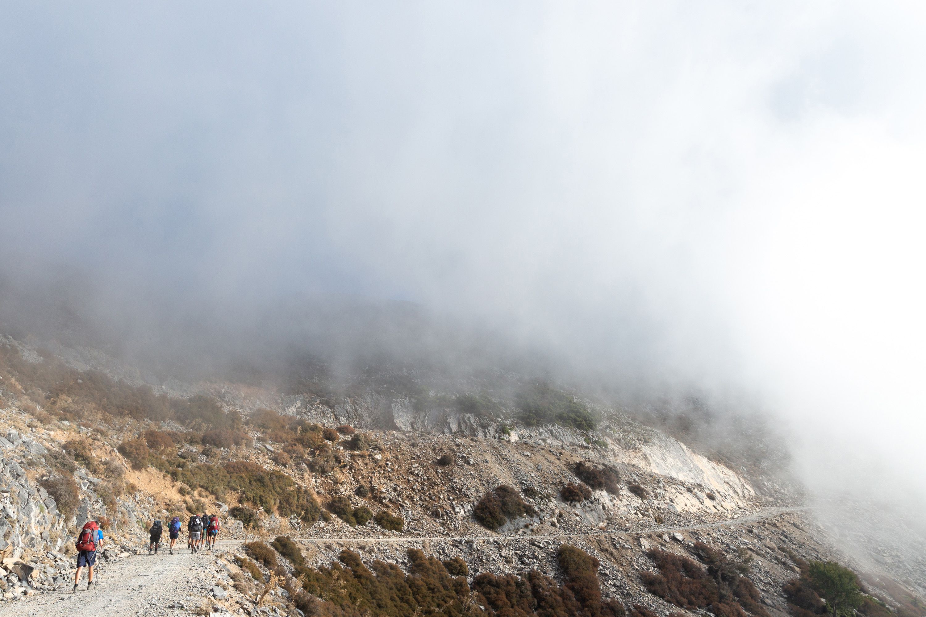 Students hiking along the side of a scree-strewn mountain in Crete. Mist obscures the ground above and ahead of them.
