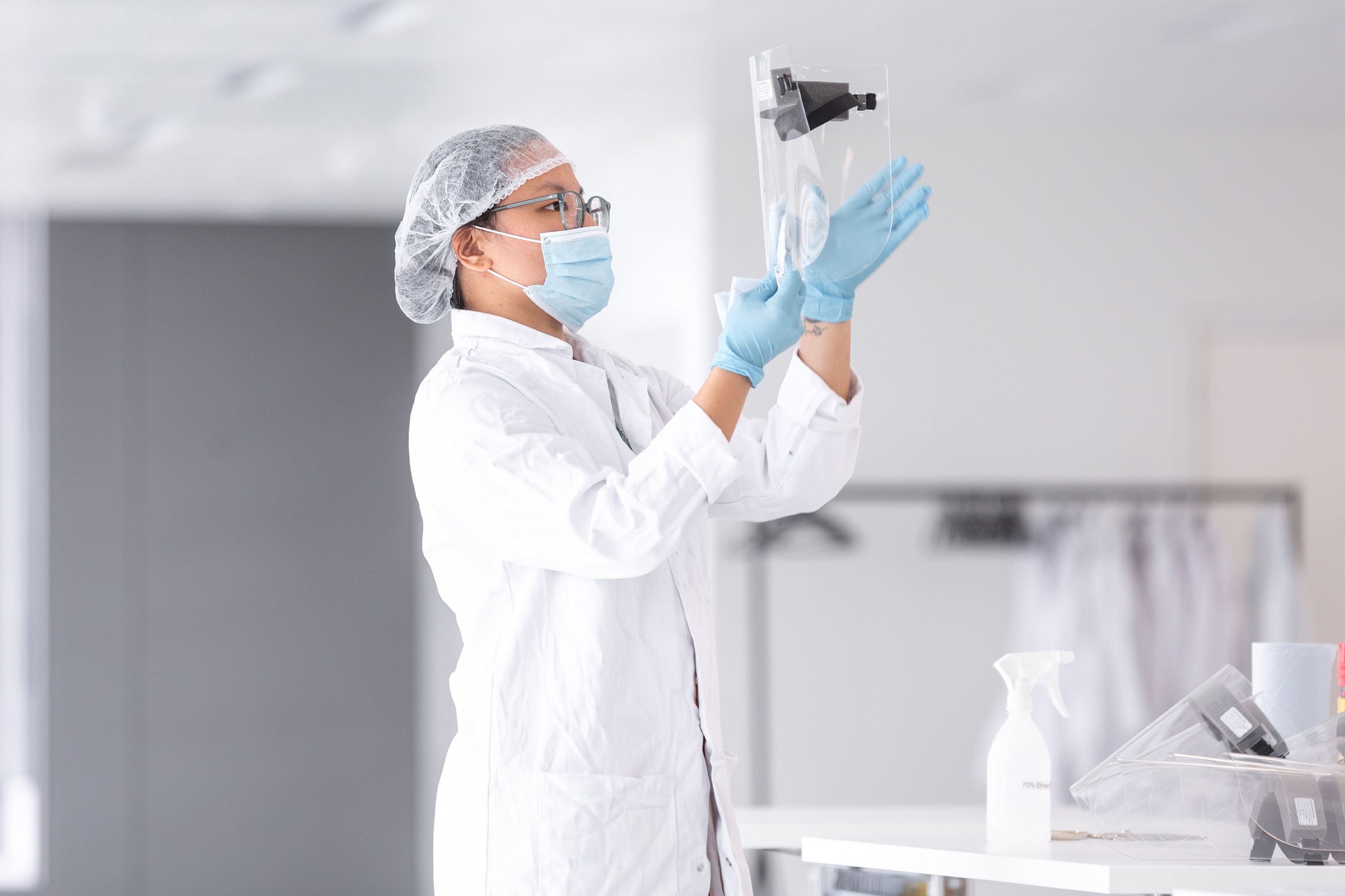 In a white room, a person in a white lab coat and hairnet examines a PPE facemask