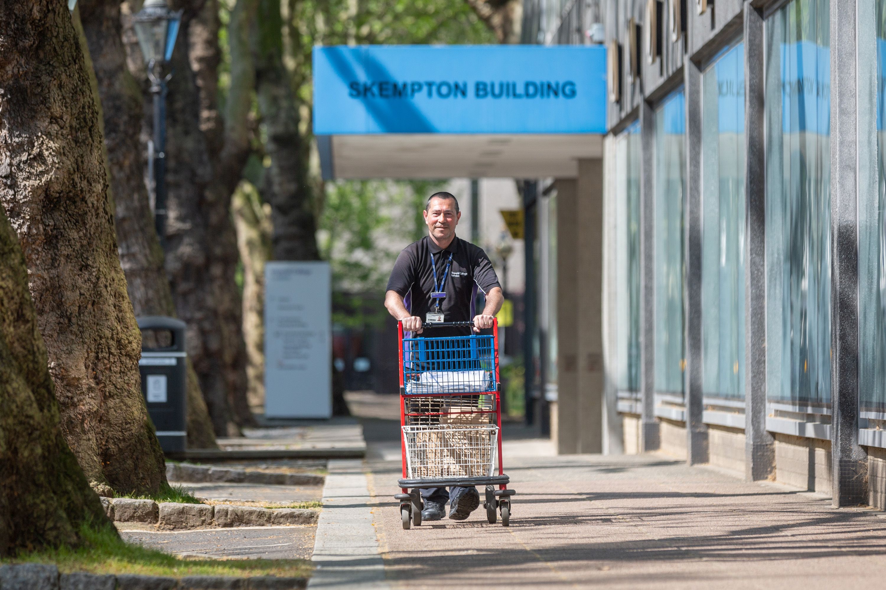 A smiling man in a blue Imperial shirt wheels a post trolley outside the Skempton Building