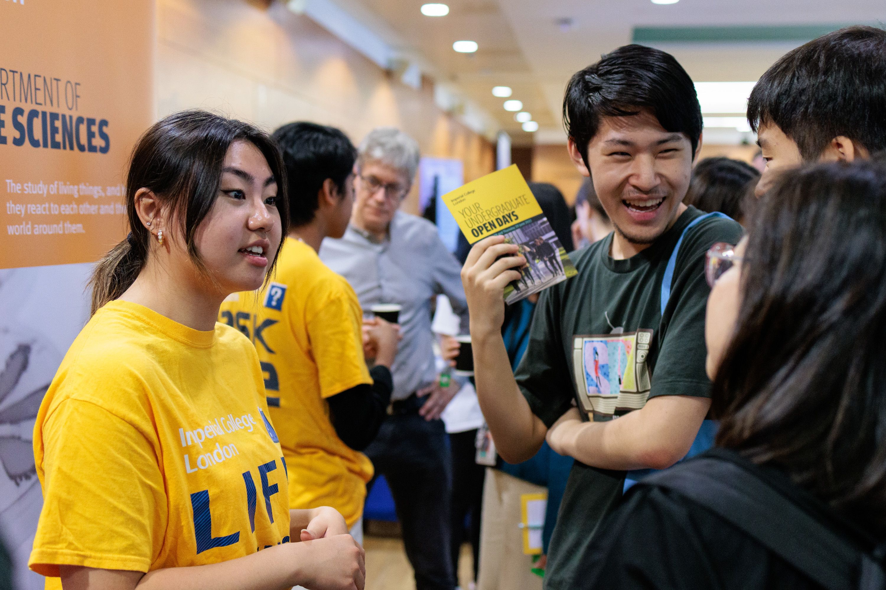 A woman in a yellow Imperial T-shirt talks to some laughing visitors