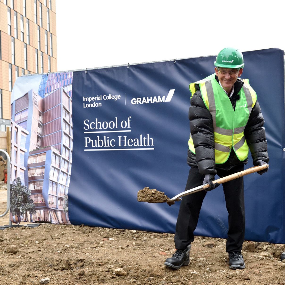 Professor Neil Alford is at the site of the new SPH building. He wears a helmet and safety equipment and holds a spadeful of soil to illustrate the idea of 'breaking ground' 