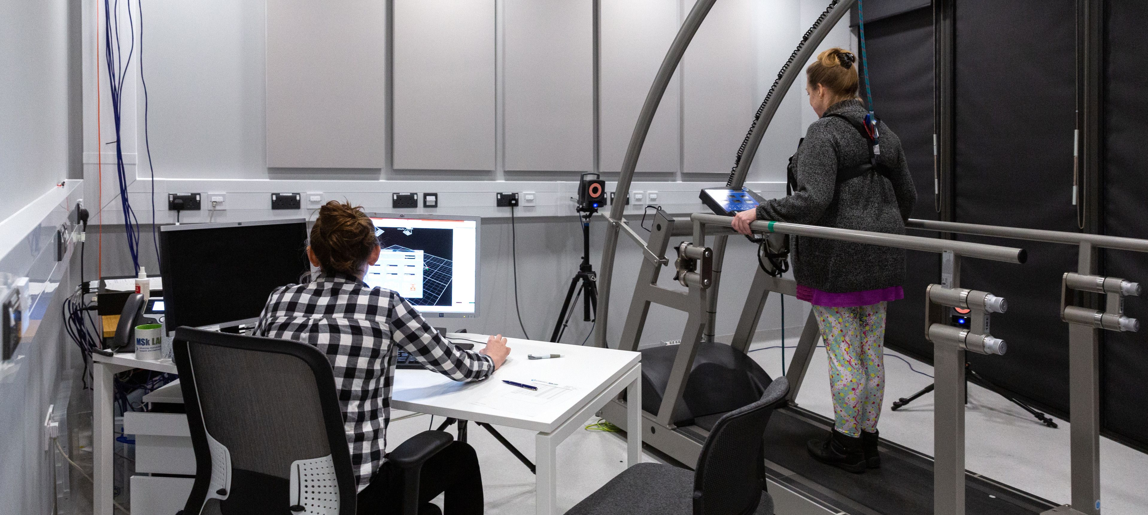 A woman at a computer observing another woman on a treadmill while attached with a harness from above