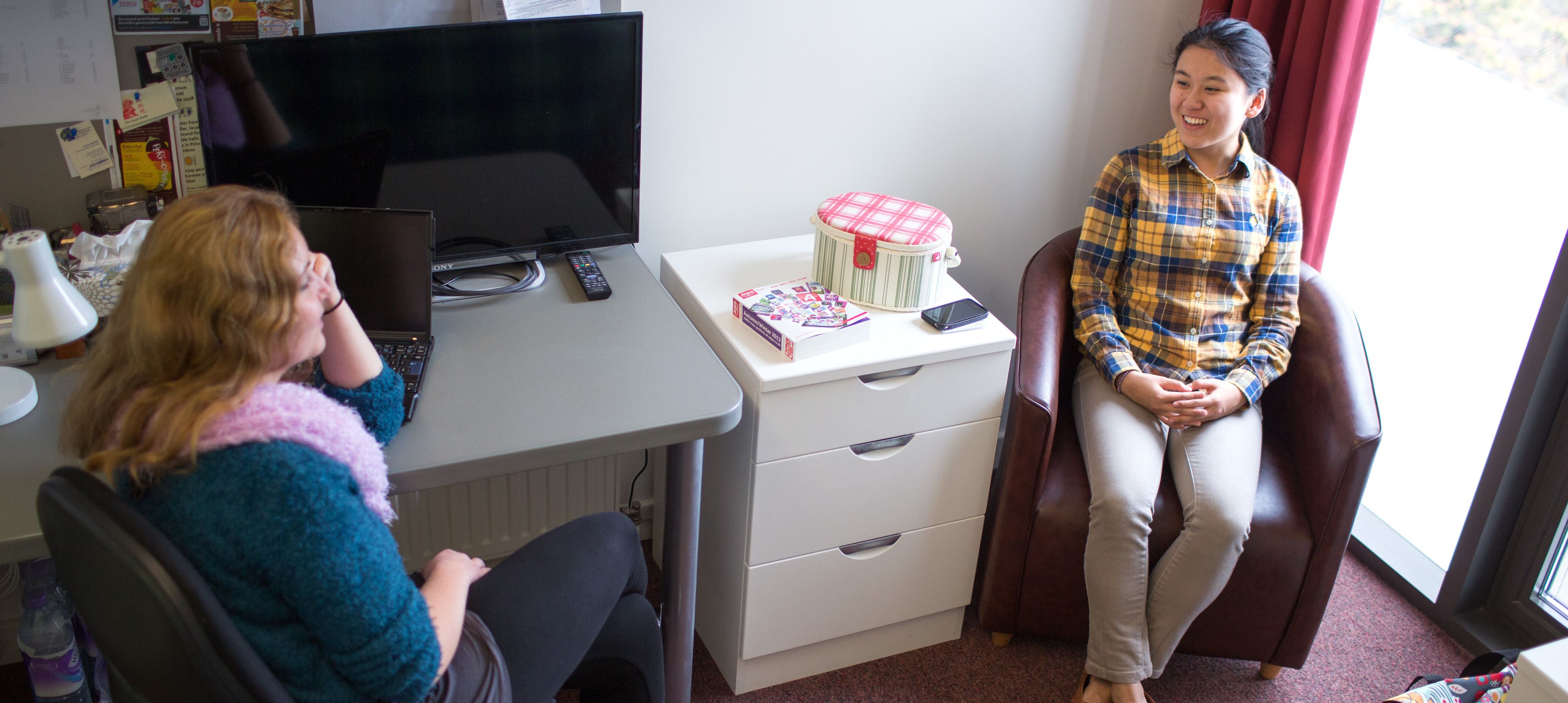 A student sitting at a desk chatting to another student who is sitting in a brown tub chair in a study bedroom in an Imperial College London hall of residence