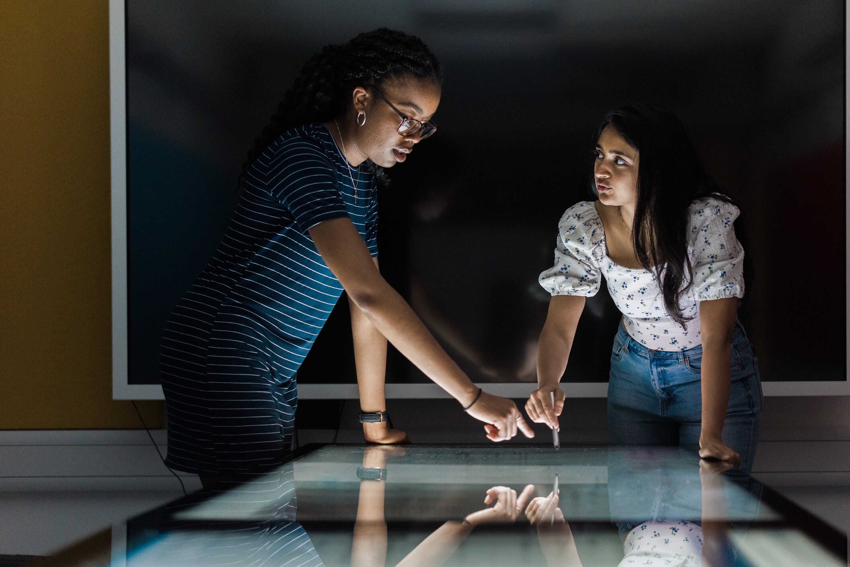 two female students in a dark room pointing at a light board