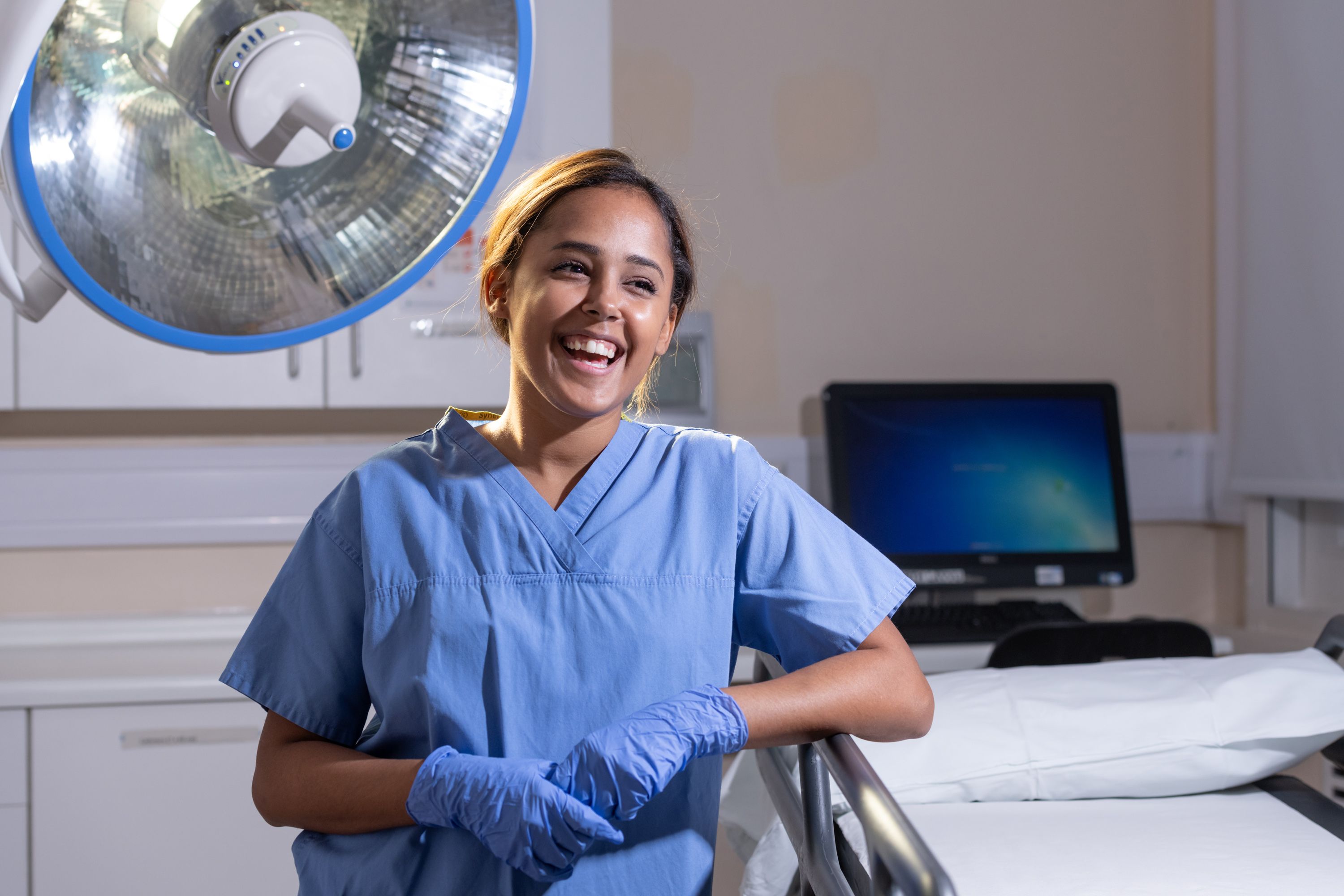 A portrait photograph of Layla Bolton Saghdaoui, who is wearing blue scrubs as she stands next to an operating theatre light