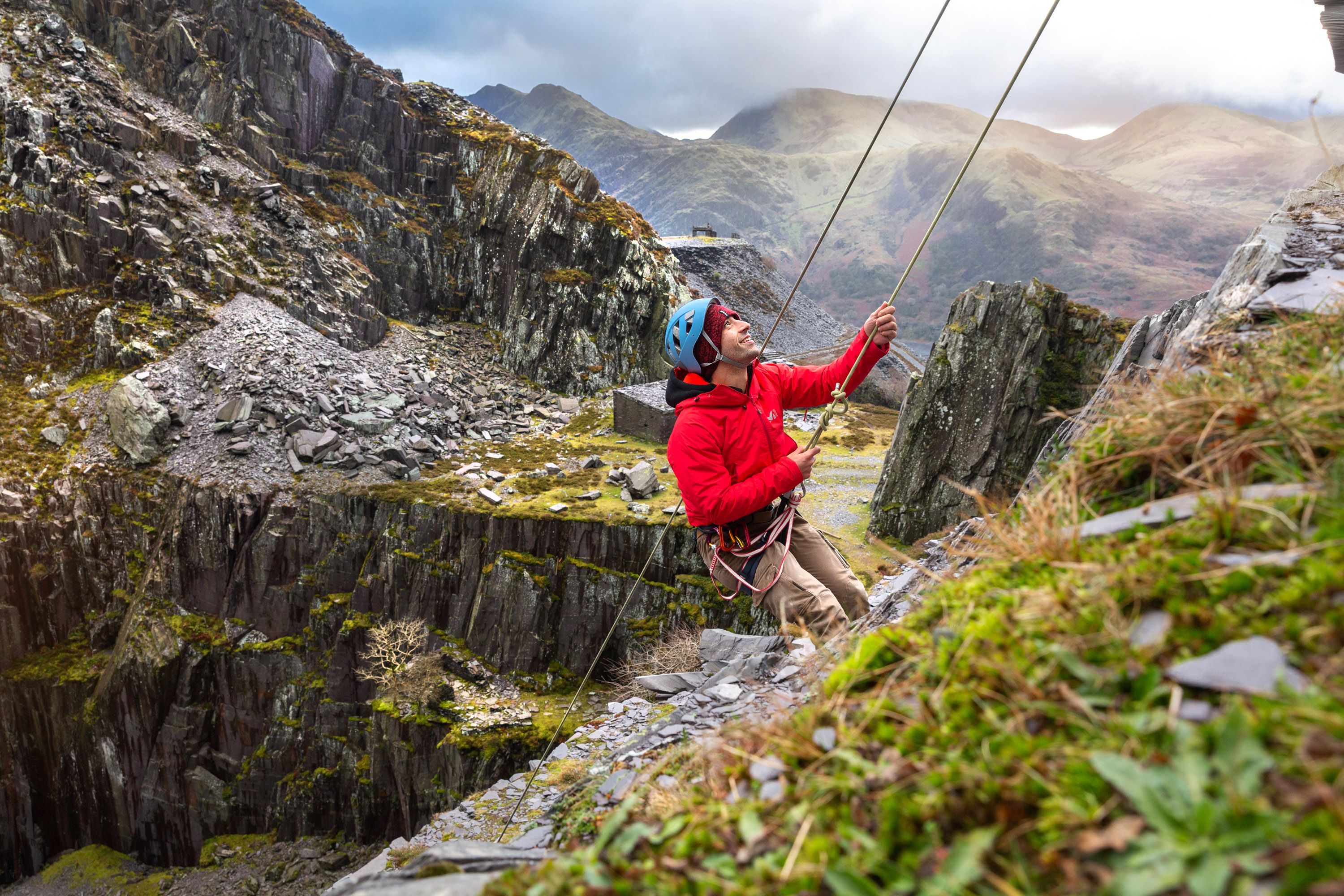 A man in a bright red jacket climbing ropes up the side of a cliff, with a dramatic landscape in the background