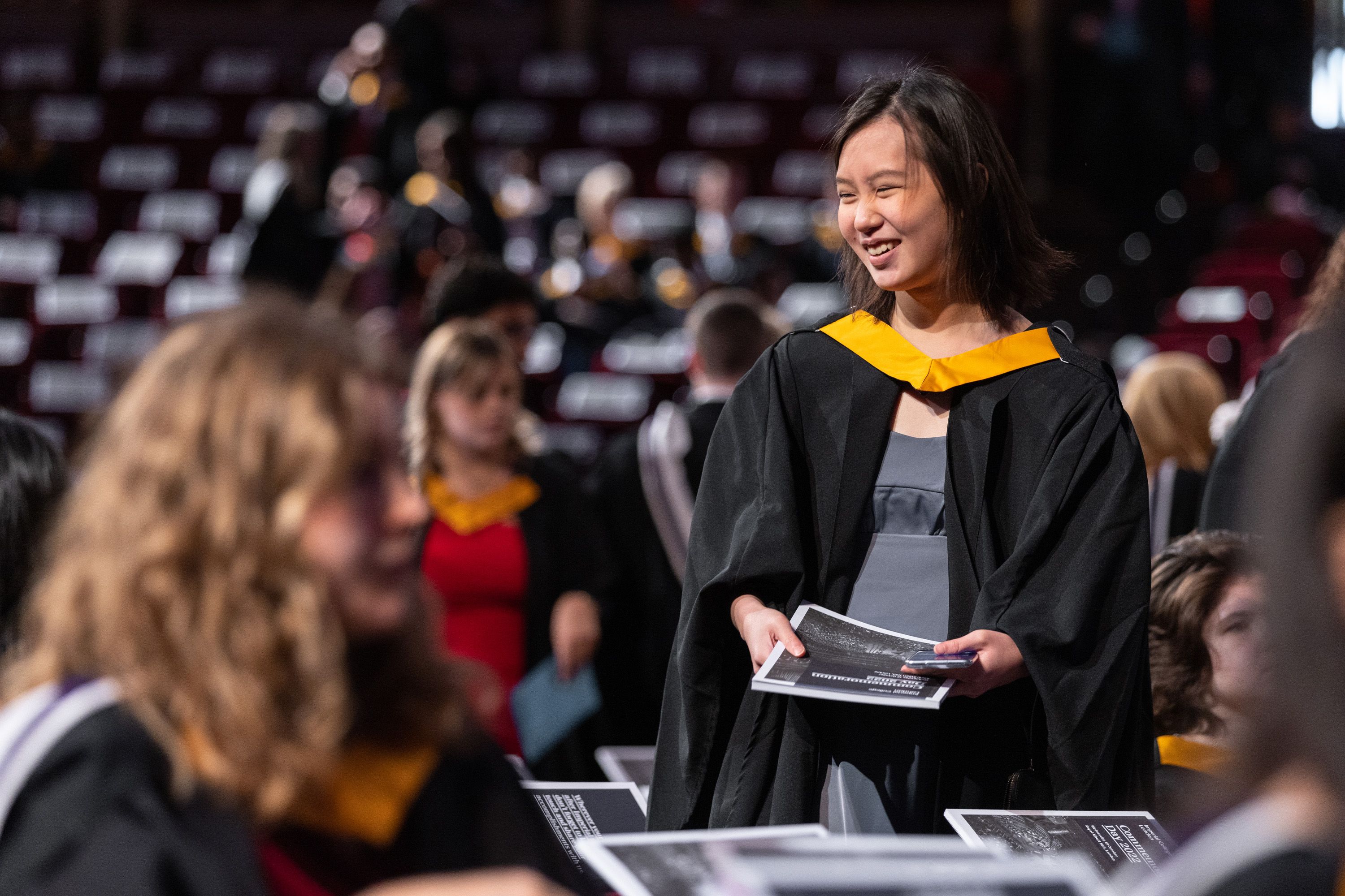 A female student in a graduation gown smiling as she holds a programme