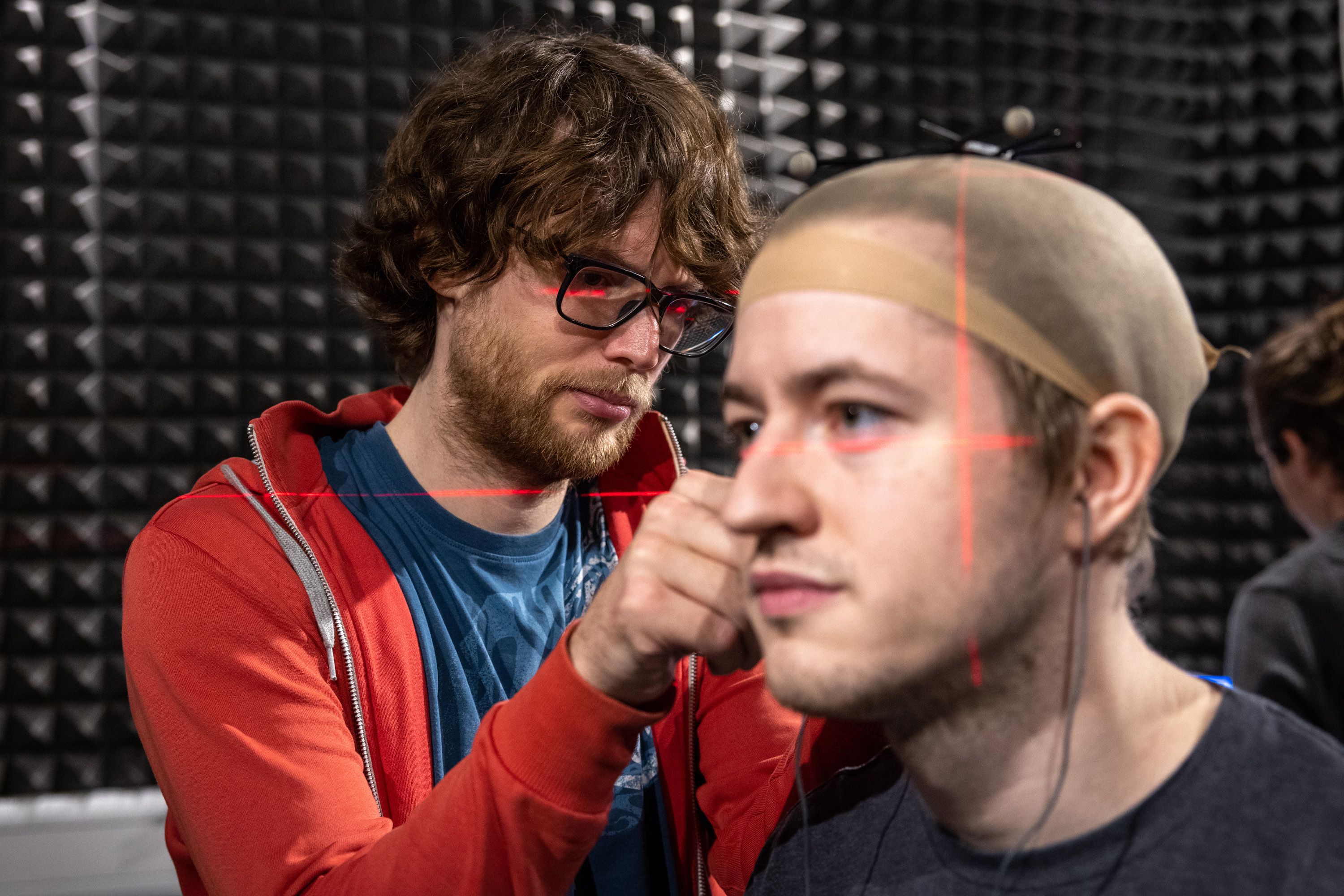 A man wearing a hairnet having equipment fitted to his ear, with red laser lines across his face