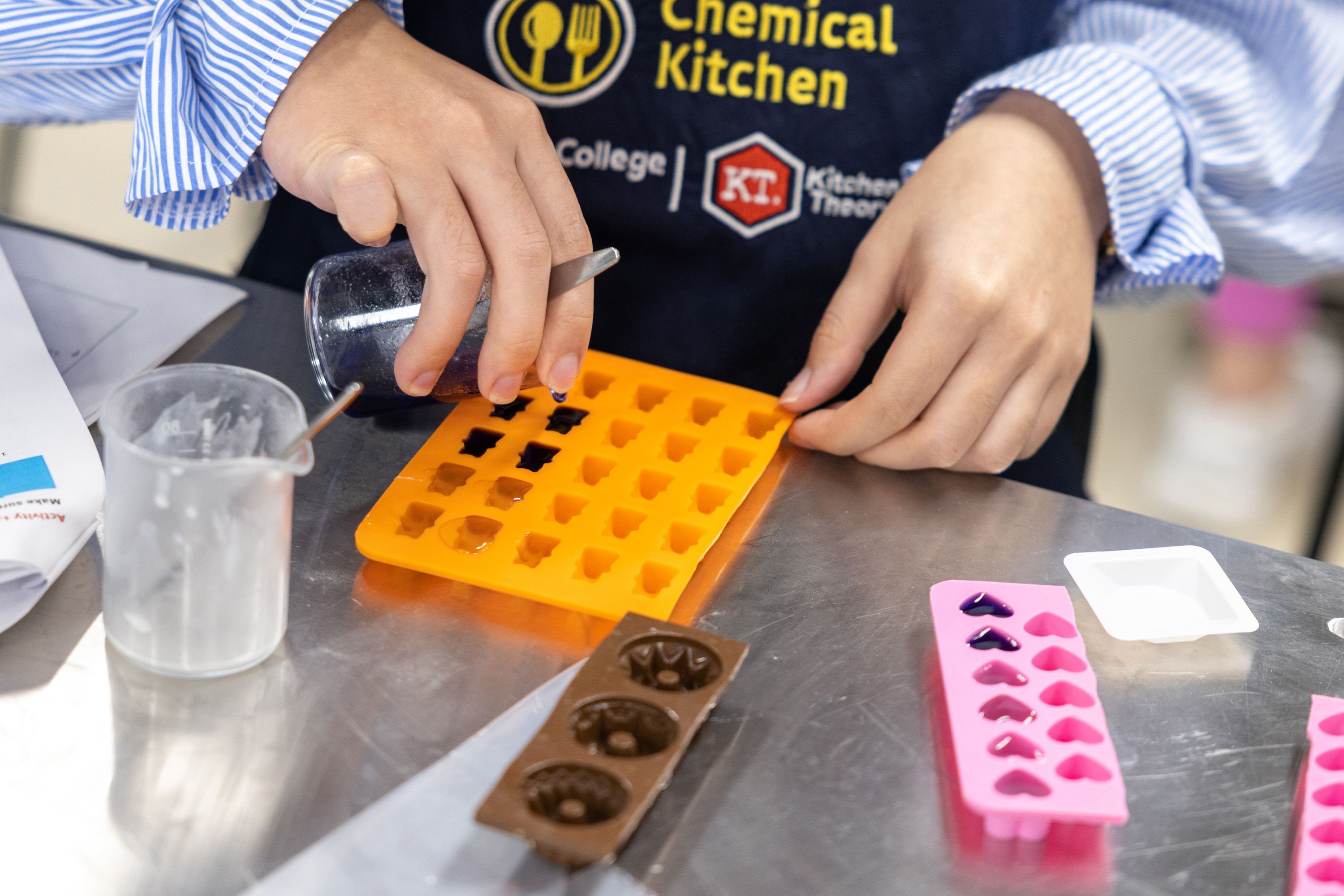 A close-up of a student's hands as they fill orange and pink pattern moulds with a dark substance