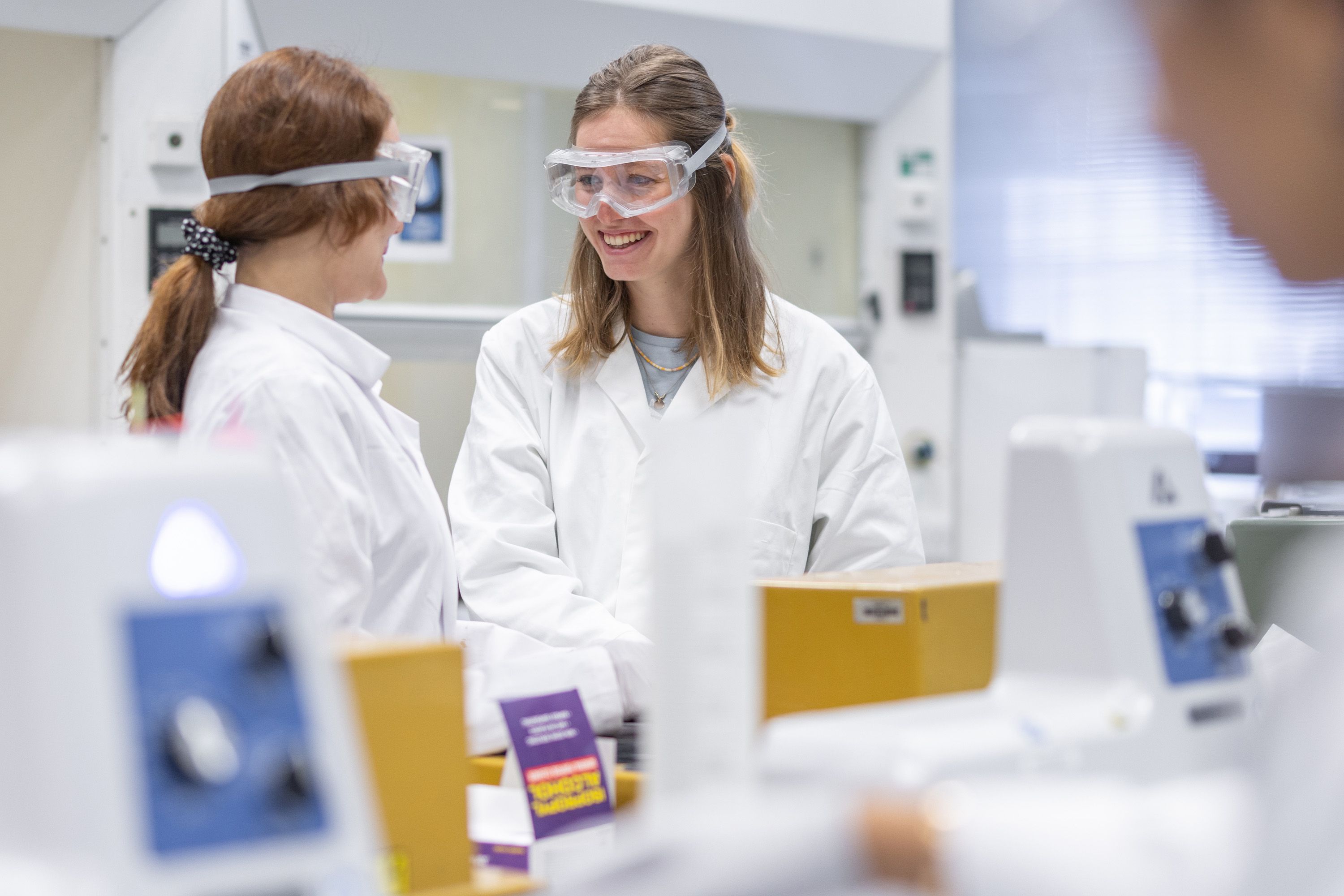 Two students in a lab wearing lab coats and eye protectors