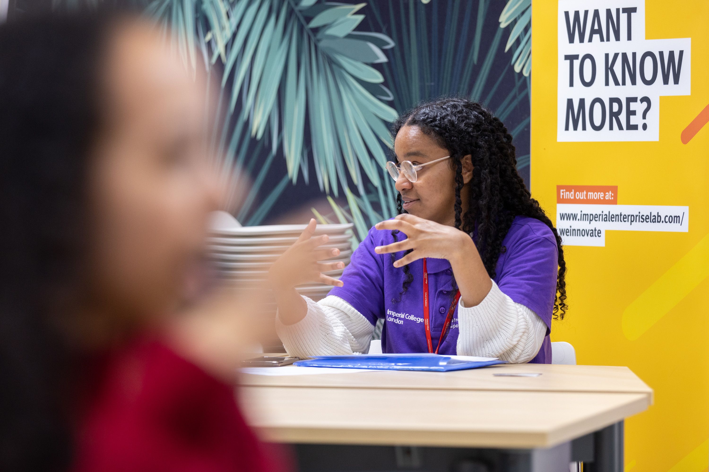 A woman in a purple Imperial top sits at a table talking to students