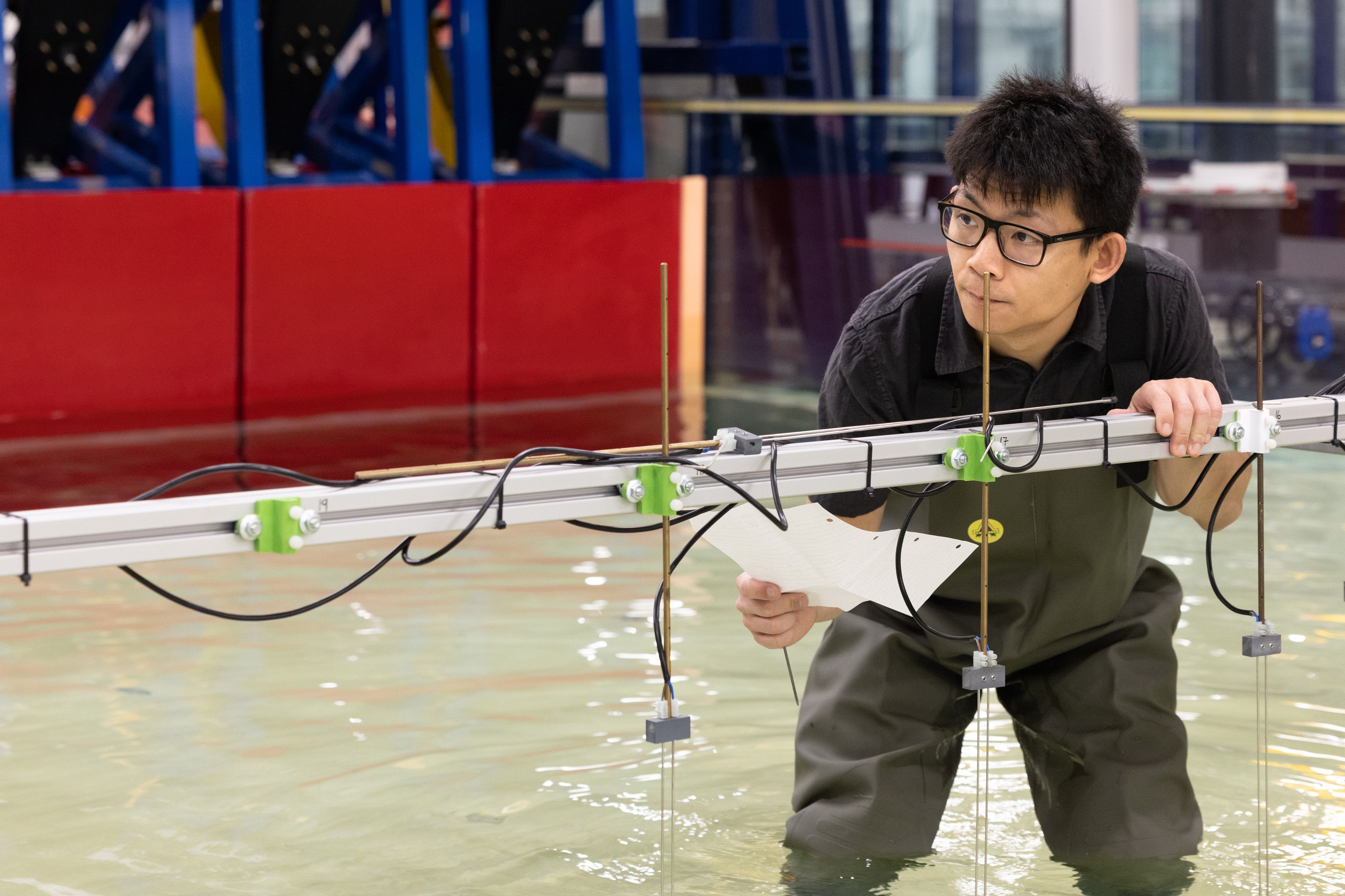 A man standing up to his knees in water in the wave tank, examining the equipment