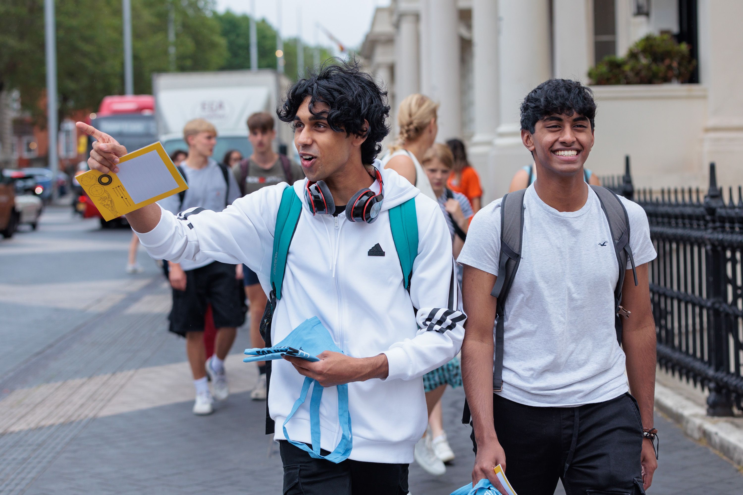 Two young men wearing backpacks walking down Exhibition Road smiling, one of them pointing off camera