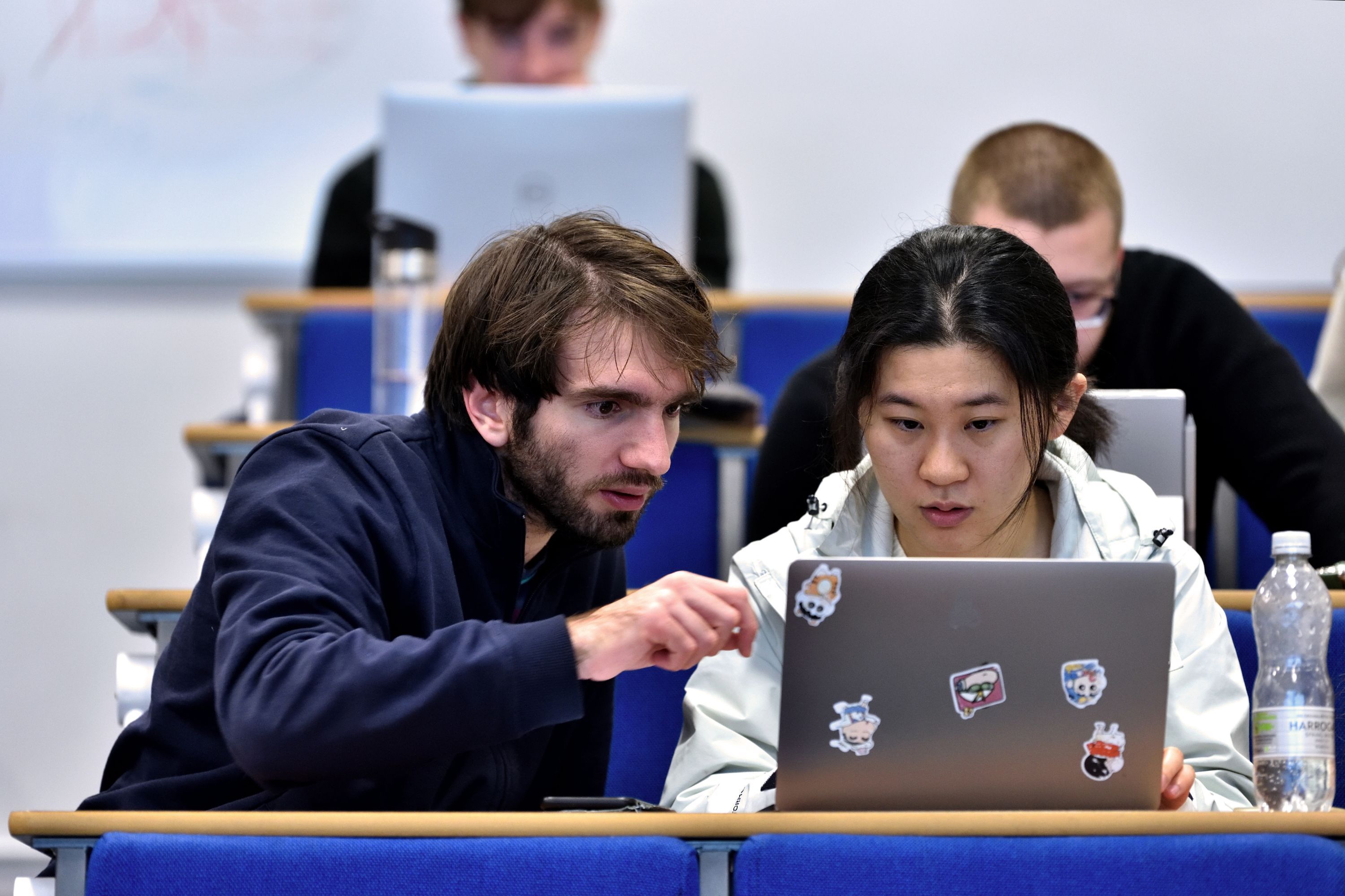 two students in a lecture room sitting at a desk looking at a laptop and discussing