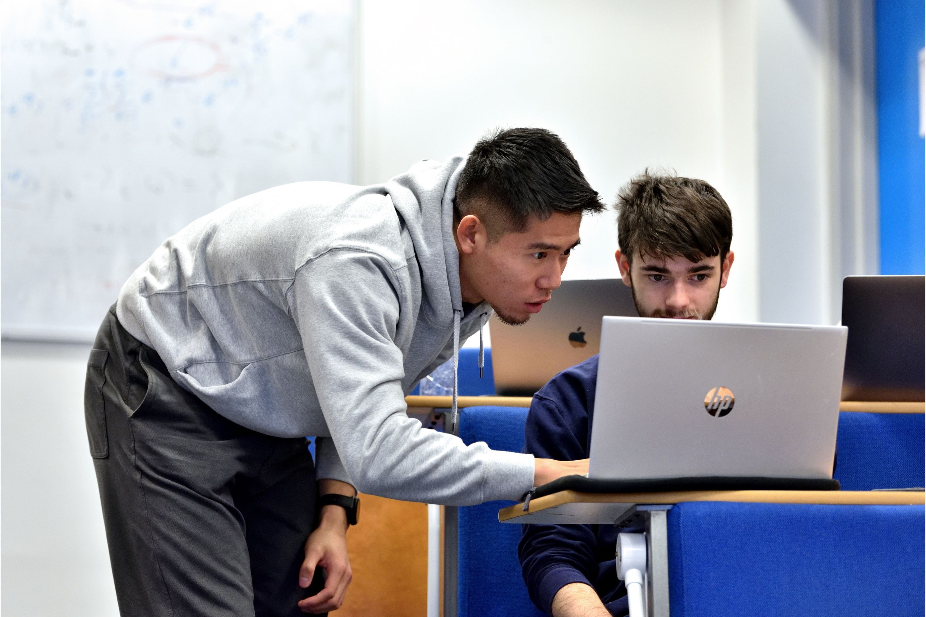 two students in a lecture room sitting at a desk looking at a laptop and discussing