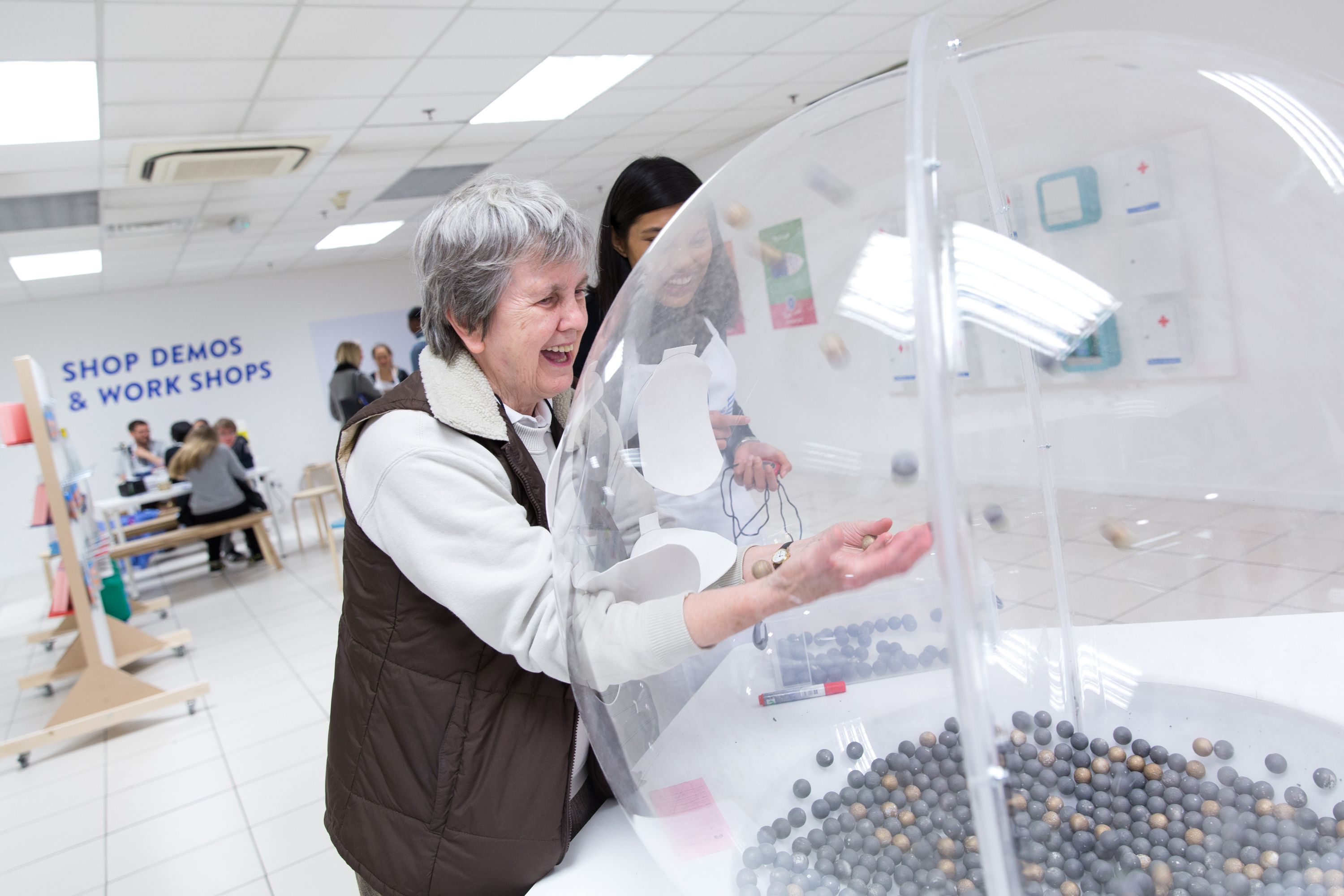 A laughing woman puts her arms inside a huge transparent plastic globe which has a layer of grey balls at the bottom