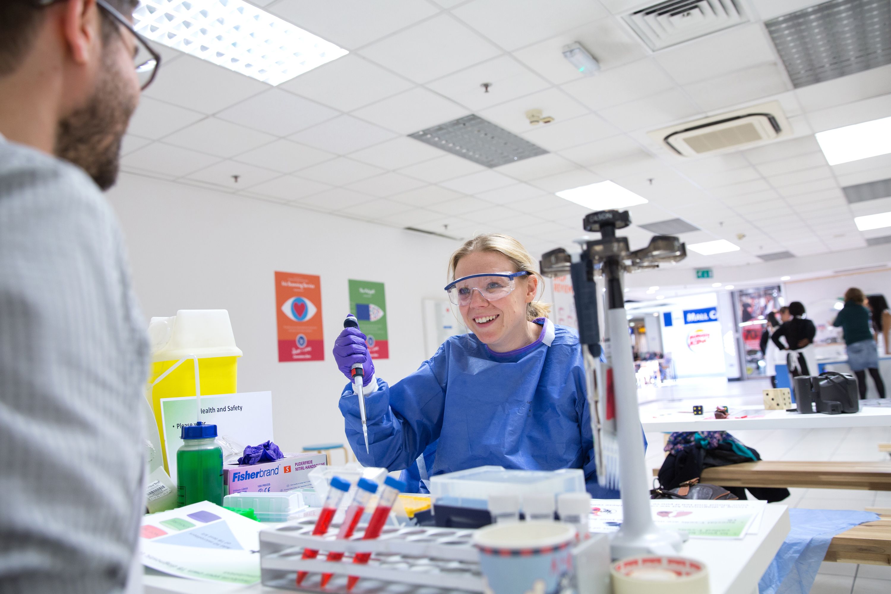 A smiling woman in blue scrubs and holding a piece of medical equipment sits at a table opposite a man in a while shirt