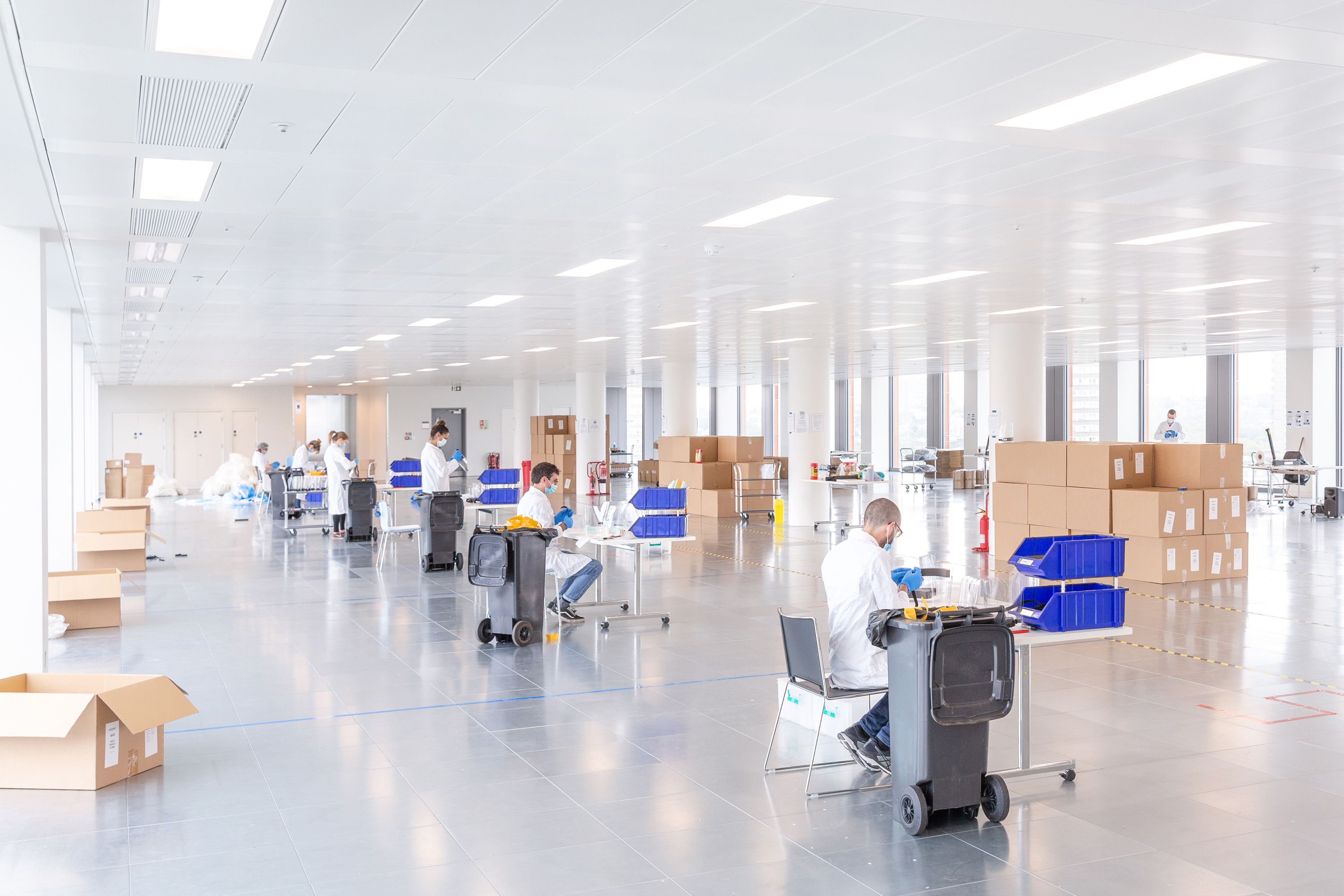 In a huge white room, people in lab coats at widely spaced tables work creating PPE face masks